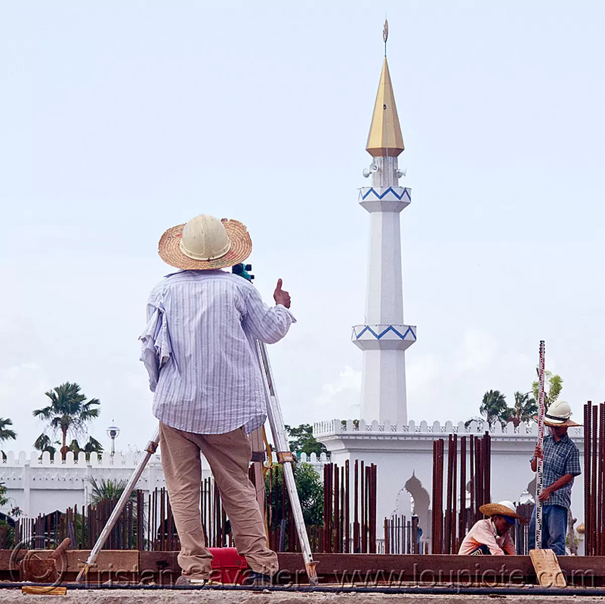 construction surveying - theodolite and tower ruler, building construction, construction site, construction surveyor, construction workers, geometer, man, minaret, miri, mosque, safety helmet, standing, straw hat, sun hat, survey, thumb up, tripod, stock photo