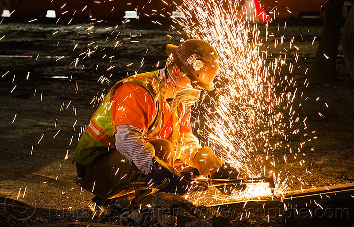 welder cutting a rail with an oxy-acetylene torch, dust mask, light rail, man, muni, night, ntk, oxy-acetylene cutting torch, oxy-fuel cutting, railroad construction, railroad tracks, rails, reflective vest, safety glasses, safety helmet, safety vest, san francisco municipal railway, sparks, track maintenance, track work, worker, stock photo