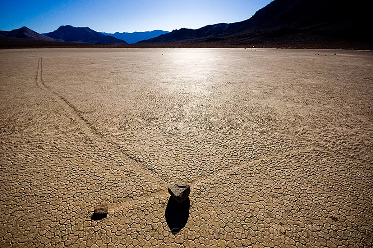 sliding rocks on the racetrack (death valley), backlight, cracked mud, crossing, desert, dry lake, dry mud, mountains, moving rocks, racetrack playa, sailing stones, tracks, stock photo