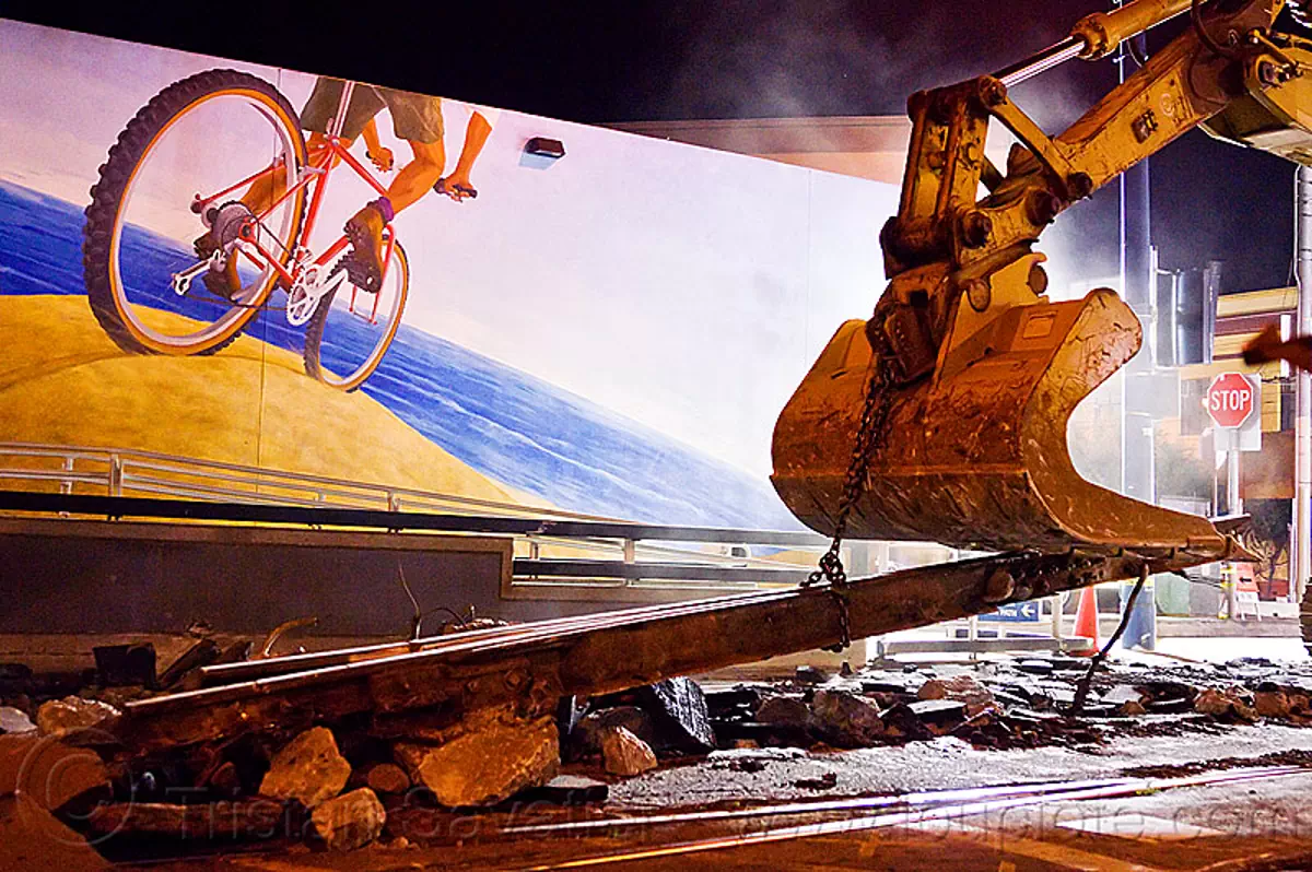 demolition of muni tracks in front of the bike mural at  church and duboce, bicycle mural, bucket attachment, caterpillar, chain, excavator, light rail, machinery, night, ntk, railroad construction, railroad tracks, rails, san francisco municipal railway, track maintenance, track work, stock photo