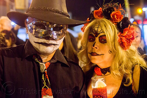 man with black mosaic mask and hat - dia de los muertos (san francisco), black hat, carnival mask, day of the dead, dia de los muertos, face painting, facepaint, flower headdress, flowers, glass candle, halloween, man, mosaic mask, night, red, skull makeup, woman