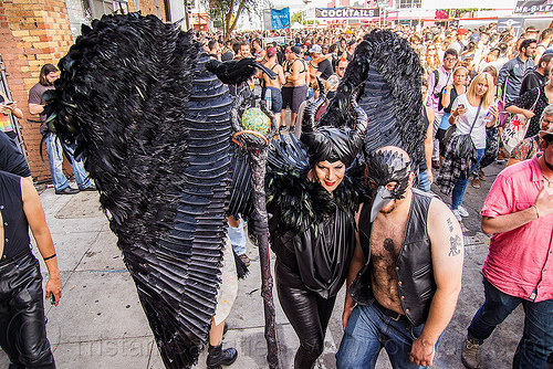 woman in black angel costume, angel costume, angel wings, bird mask, black angel, black bird, black feathers, ceremonial staff, ceremonial stick, crowd, man, woman