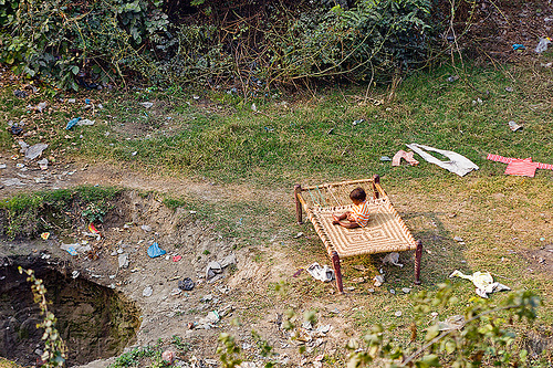 child sitting on a bed frame in a field, bed, child, cloth, drying, environment, garbage, kid, plastic trash, pollution, sink hole, sitting, toddler
