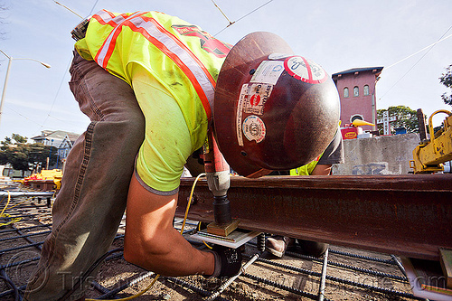 construction worker bolting rail ties, bolts, compact gun, construction worker, duboce, high-visibility jacket, high-visibility vest, light rail, man, muni, ntk, power tool, railroad construction, railroad ties, railroad tracks, railway tracks, reflective jacket, reflective vest, safety helmet, safety vest, san francisco municipal railway, track maintenance, track work, working