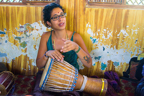 maryam with djembe drum, arm tattoo, bindi, bird tattoo, bracelets, djembe drum, drummer, eyeglasses, eyewear, maryam, musical instrument, necklaces, peeling paint, percussion, phoenix tattoo, prescription glasses, rishikesh, sitting, spectacles, tattoos, woman