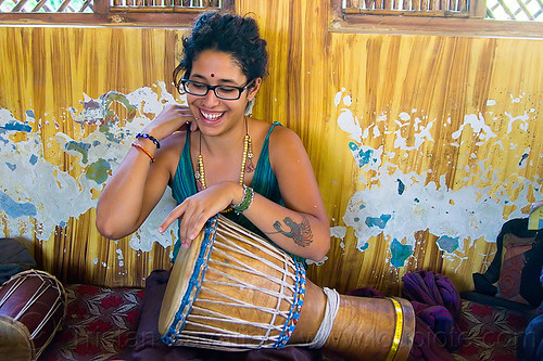 maryam with djembe drum, arm tattoo, bindi, bird tattoo, bracelets, djembe drum, drummer, eyeglasses, eyewear, maryam, musical instrument, necklaces, peeling paint, percussion, phoenix tattoo, prescription glasses, rishikesh, sitting, spectacles, tattoos, woman