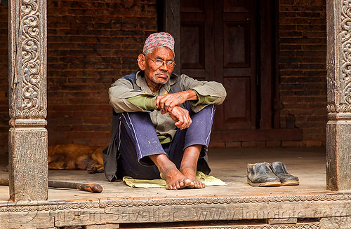 old nepali man sitting in a pati (nepal), bare feet, bhaktapur, hat, old man, pati, pillars, prescription glasses, shoes, sitting, spectacles, walking cane, walking stick, wood carving, wooden