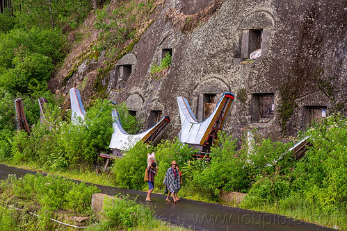 toraja people walking in front of toraja rock-tombs, burial site, cemetery, cliff, graves, graveyard, liang pak, miniature tongkonan, road, rock tombs, tana toraja, tongkonan roof, walking