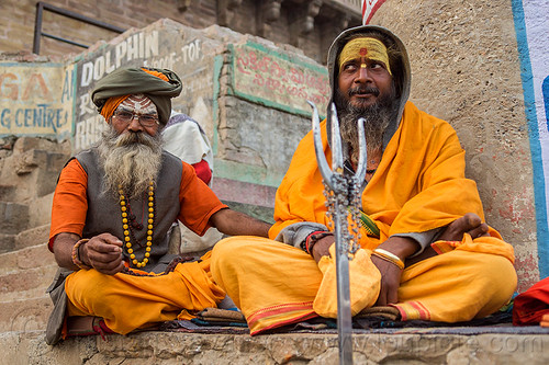 two sadhus with trident on the ghats of varanasi - hindu holy men (india), babas, beard, bhagwa, cross-legged, ghats, headdress, hindu, hinduism, men, sadhus, saffron color, sitting, tilak, tilaka, trident, turban, varanasi