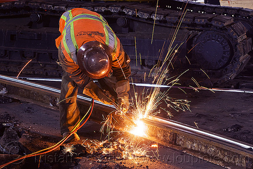 welder cutting a railroad track rail with an oxy-acetylene torch, demolition, high-visibility jacket, high-visibility vest, light rail, man, muni, night, ntk, oxy-acetylene cutting torch, oxy-fuel cutting, railroad construction, railroad tracks, railway tracks, reflective jacket, reflective vest, safety helmet, safety vest, san francisco municipal railway, sparks, track maintenance, track work, welder, welding, worker, working