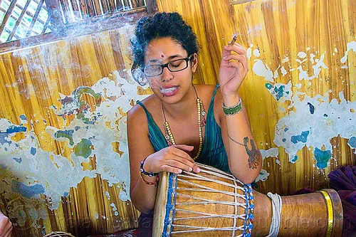 maryam smoking a joint, arm tattoo, bindi, bird tattoo, bracelets, cigarette, djembe drum, drummer, eyeglasses, eyewear, ganja, joint, maryam, musical instrument, necklaces, peeling paint, percussion, phoenix tattoo, prescription glasses, rishikesh, sitting, smoke, smoking, spectacles, tattoos, weed, woman