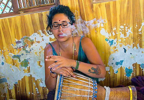 maryam smoking a joint, arm tattoo, bindi, bird tattoo, bracelets, cigarette, djembe drum, drummer, eyeglasses, eyewear, ganja, joint, maryam, musical instrument, necklaces, peeling paint, percussion, phoenix tattoo, prescription glasses, rishikesh, sitting, smoke, smoking, spectacles, tattoos, weed, woman