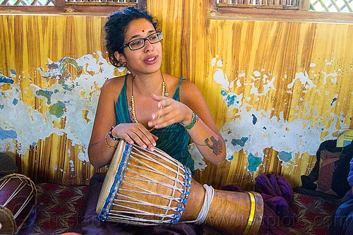 maryam with djembe drum, arm tattoo, bindi, bird tattoo, bracelets, djembe drum, drummer, eyeglasses, eyewear, maryam, musical instrument, necklaces, peeling paint, percussion, phoenix tattoo, prescription glasses, rishikesh, sitting, spectacles, tattoos, woman