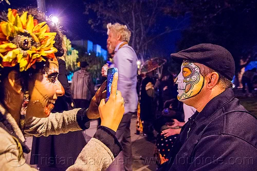 woman taking cellphone photo of man with sugar skull face paint - dia de los muertos, camera, cellphone, day of the dead, dia de los muertos, face painting, facepaint, halloween, hat, man, night, photographer, sahar, sugar skull makeup, taking photo, taking picture, woman
